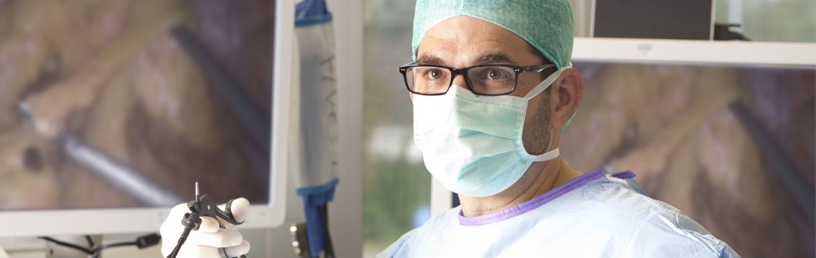 Ein Arzt steht im Op und führt ein Werkszeug bei einem minimalinvasiven Eingriff. Neben und hinter ihm sind große Bildschirme zu sehen auf denen das Operationsgebiet zu sehen ist.