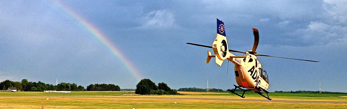 Ein gelber Rettungshubschrauber vom ADAC hebt von einer grünen Wiese ab. Im Hintergrund stehen viele Bäume und ein Regenbogen ist zu sehen.