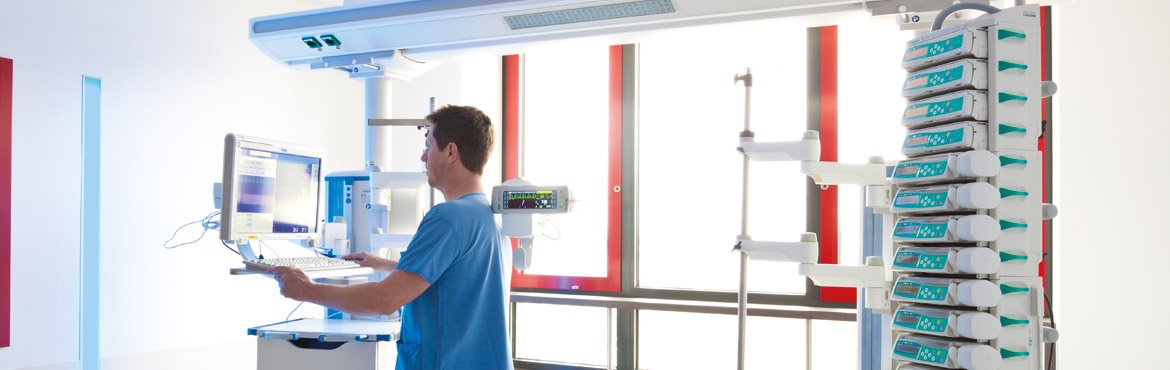 Ein Pfleger steht vor einem Computermonitor in einer leeren Patientennische auf der Intensivstation. Vor ihm und hinter ihm sind medizinische Geräte zu sehen.