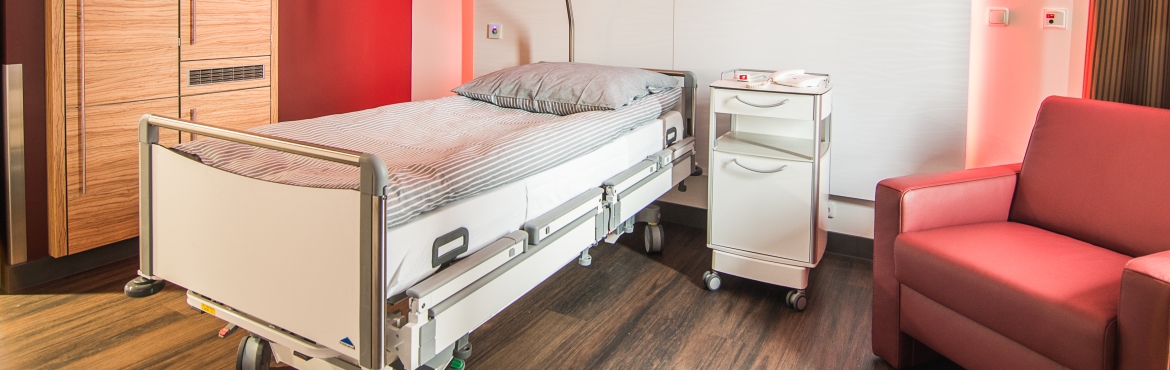Blick vom Fenster in ein Patientenzimmer auf ein weißes Krankenbett mit weiß grau gestreifter Bettwäsche. Rechts daneben ein weißer Nachtschrank auf Rollen und ein roter Sessel. Links daneben ein Holzkleiderschrank