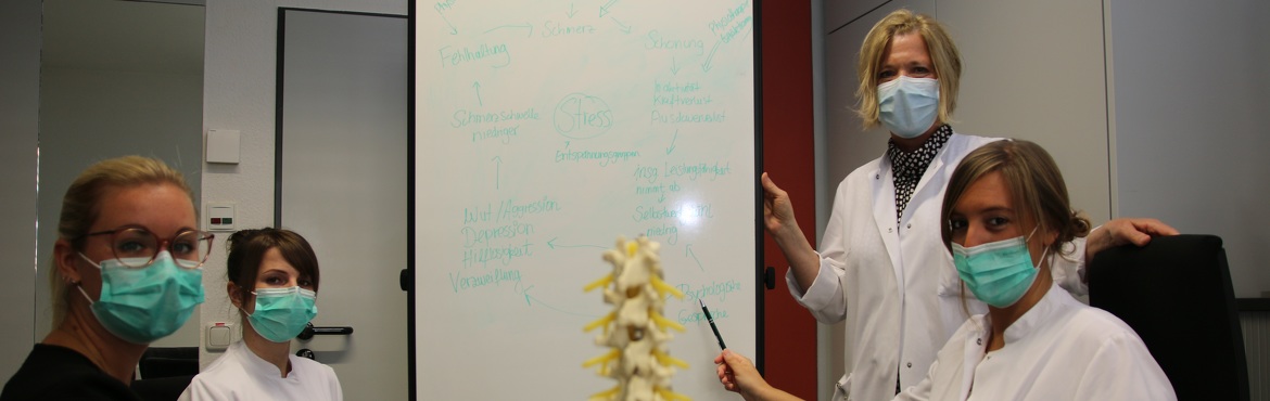 Patientin, zwei Psychologinnen und eine Ärztin schauen in die Kamera und unterhalten sich. Alle tragen einen Mund-Nasen-Schutz. In der Mitte steht ein Modell einer Wirbelsäule und eine Flippchart.