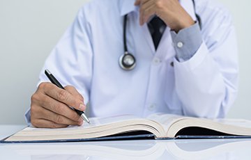 Ein Arzt im weißen Kittel sitzt an einem weißen Tisch. Er trägt ein Stethoskop um den Hals und hält einen Stift in der Hand. Vor ihm liegt ein großes aufgeschlagenes Buch auf einem weißen Tisch.