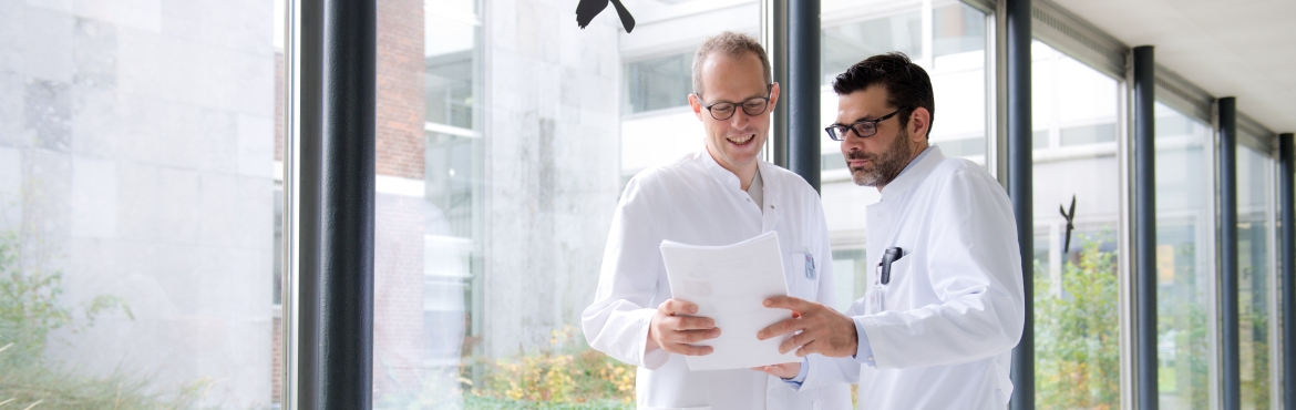 Zwei Ärzte stehen in einem hellen Gang des Krankenhauses. Biede haben einen weißen Kittel an und haben Unterlagen in der Hand auf die sie schauen.