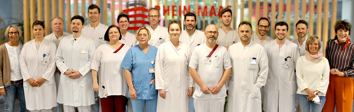 Achtzen Ärztinnen und Ärzte und zwei Sekretärinnen von der Klinik für Allgemein-, Viszeral- und Minimalinvasive Chirurgie stehen vor einer beigen Wand im Rhein-Maas Klinikum. Die Ärztinnen und Ärzte tragen einen weißen Kittel und eine weiße Hose.