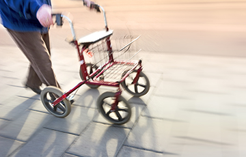 Ein Mann, gekleidet mit einer braunen Hose und einer blauen Jacke, geht mit seinem Rollator über einen Gehweg.