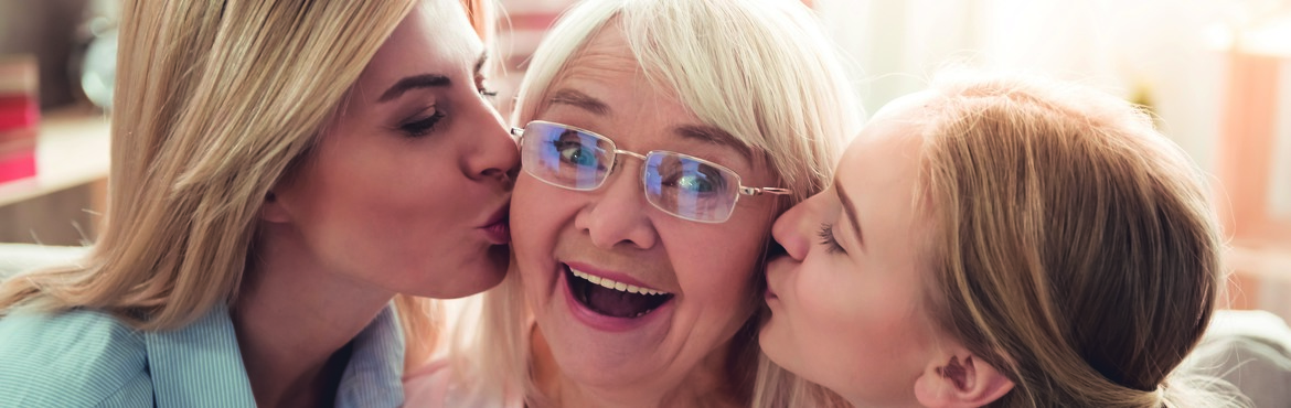 Eine ältere Frau wird von einer jüngeren Frau und einem jungen Mädchen rechts und links auf die Wange geküsst und lacht dabei